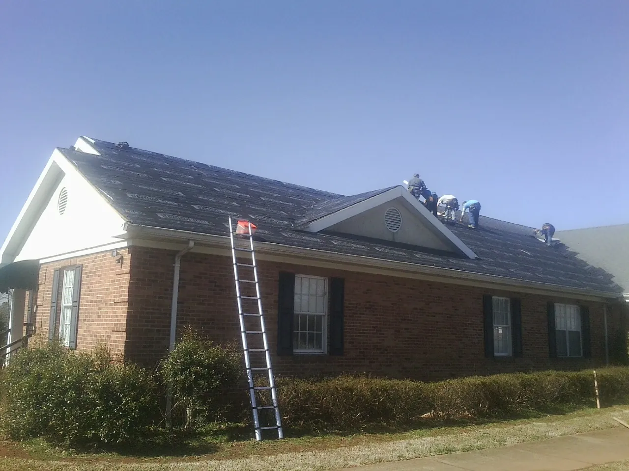 Crew tearing shingles off a brick ranch roof with a ladder set — Emergency Roof Repair work by our crew in Cochrane