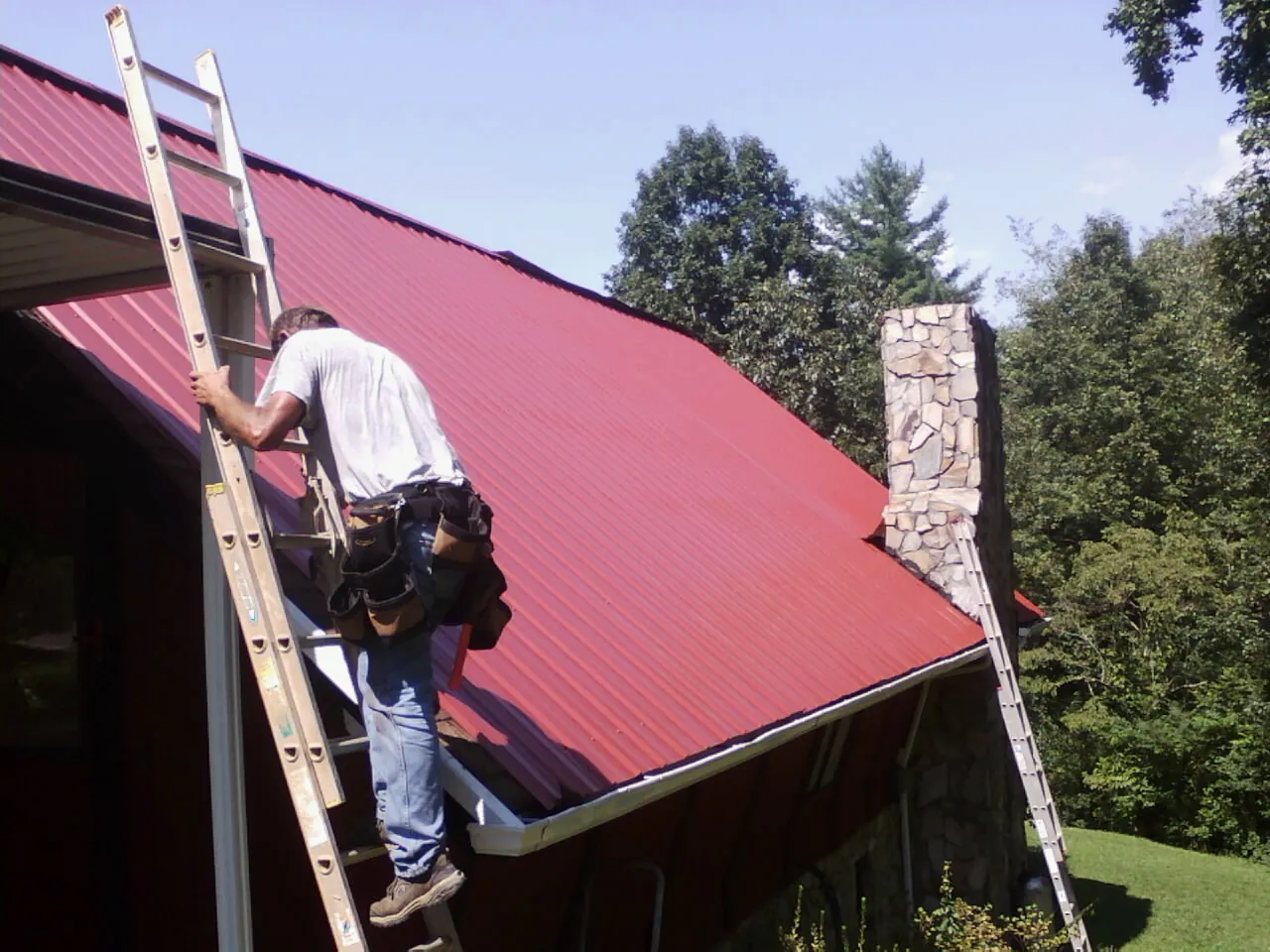 Roofer on a ladder beside a red metal roof and stone chimney — Soffit & Fascia Repair by Roofing Brothers in Cochrane